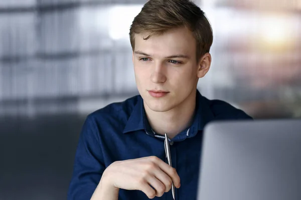 Jeune homme d'affaires blond réfléchissant à la stratégie sur son lieu de travail avec ordinateur dans un bureau sombre, éblouissement de la lumière sur le fond — Photo