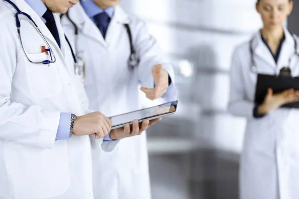 Group of unknown doctors use a computer tablet to check up some medical names records, while standing in a hospital office. Physicians ready to examine and help patients. Medical help, insurance in