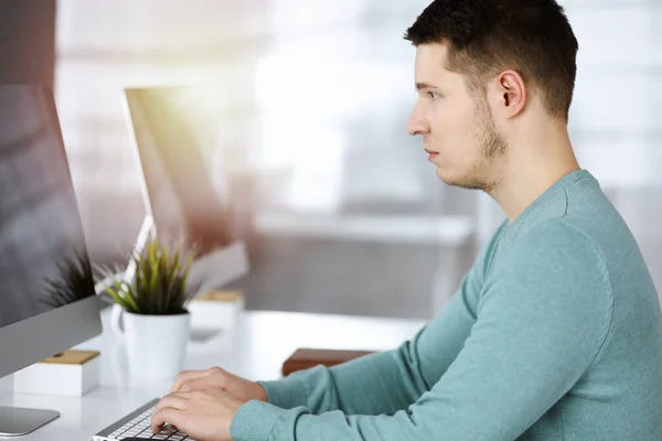 Der junge selbstbewusste Geschäftsmann sitzt in einem grünen Pullover am Schreibtisch in einem sonnigen modernen Büro, während er am Computer an seinem neuen Projekt arbeitet. Kopfschuss eines männlichen Unternehmers oder IT-Spezialisten — Stockfoto