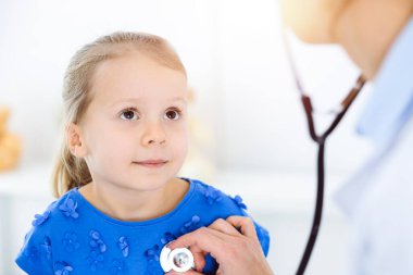 Güneşli klinikte bir çocuğu steteskopla muayene eden bir doktor. Mavi elbiseli mutlu gülümseyen kız hasta her zamanki sağlık kontrolünde.