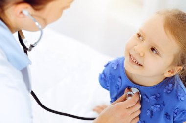 Güneşli klinikte bir çocuğu steteskopla muayene eden bir doktor. Mavi elbiseli mutlu gülümseyen kız hasta her zamanki sağlık kontrolünde.