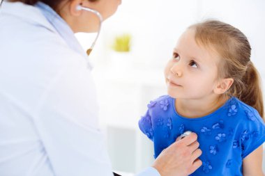 Güneşli klinikte bir çocuğu steteskopla muayene eden bir doktor. Mavi elbiseli mutlu gülümseyen kız hasta her zamanki sağlık kontrolünde.