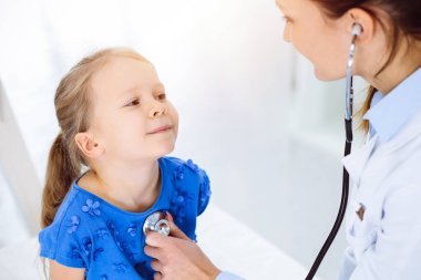 Güneşli klinikte bir çocuğu steteskopla muayene eden bir doktor. Mavi elbiseli mutlu gülümseyen kız hasta her zamanki sağlık kontrolünde.