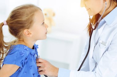 Güneşli klinikte bir çocuğu steteskopla muayene eden bir doktor. Mavi elbiseli mutlu gülümseyen kız hasta her zamanki sağlık kontrolünde.
