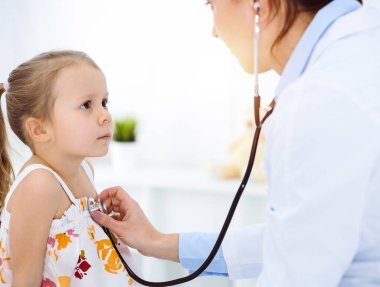 Güneşli klinikte bir çocuğu steteskopla muayene eden bir doktor. Parlak renkli elbise giymiş mutlu gülümseyen kız hasta her zamanki sağlık kontrolünde.