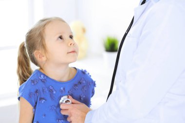 Doktor steteskopla küçük bir kızı muayene ediyor. Her zamanki tıbbi muayenede gülümseyen çocuk hasta. Tıp ve sağlık hizmetleri kavramları