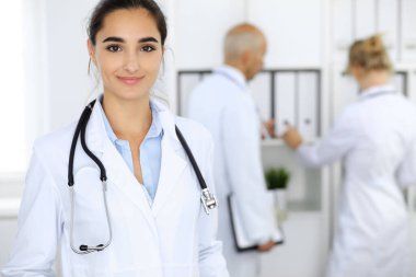 Klinikte arka planda meslektaşları olan bir kadın doktor. İspanyol ya da Latin Amerikalı tıp personeli