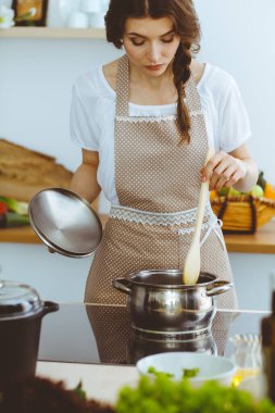 Genç esmer kadın mutfakta çorba pişiriyor. Ev hanımı elinde tahta kaşık tutuyor. Gıda ve sağlık konsepti