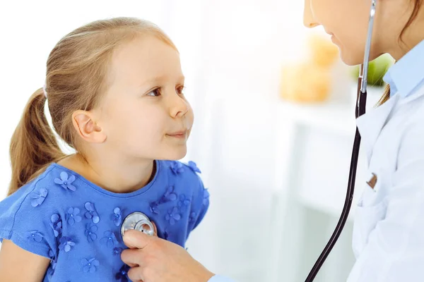 Doutor examinando uma criança por estetoscópio na clínica ensolarada. Feliz sorrindo paciente menina vestida de vestido azul é na inspeção médica habitual — Fotografia de Stock