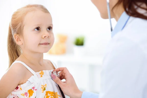 Γιατρός εξετάζει ένα παιδί με στηθοσκόπιο σε ηλιόλουστη κλινική. Ευτυχισμένο χαμογελαστό κορίτσι ασθενή ντυμένος με φωτεινό χρώμα φόρεμα είναι στη συνήθη ιατρική επιθεώρηση — Φωτογραφία Αρχείου