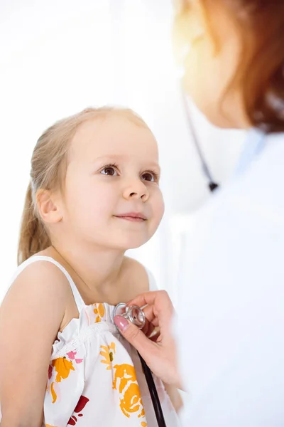 Médico examinando a un niño por estetoscopio en una clínica soleada. Feliz niña sonriente paciente vestida con vestido de color brillante es en la inspección médica habitual — Foto de Stock