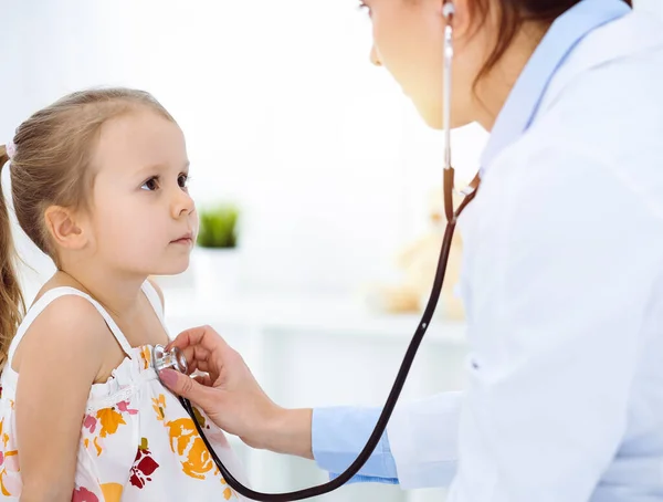 Doutor examinando uma criança por estetoscópio na clínica ensolarada. Feliz sorrindo paciente menina vestida com vestido de cor brilhante é na inspeção médica habitual — Fotografia de Stock