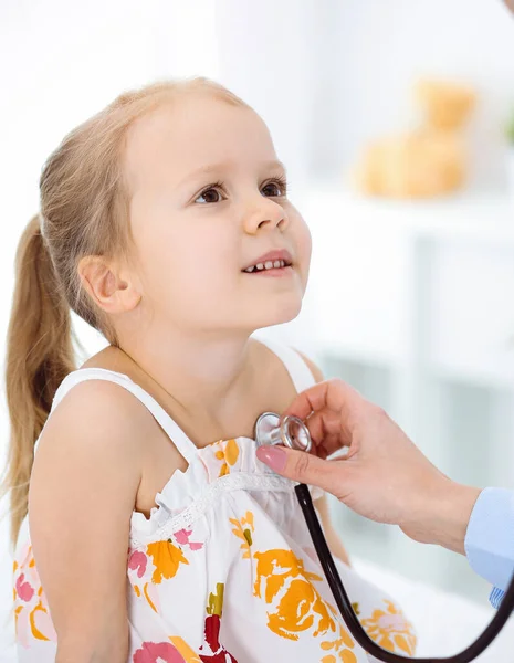 Médecin examinant un enfant par stéthoscope dans une clinique ensoleillée. Heureuse fille souriante patient habillé en robe de couleur vive est à l'inspection médicale habituelle — Photo