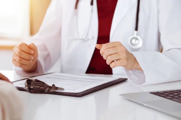 Mujer-médico desconocida y paciente discutiendo algo mientras se sienta a la mesa en la clínica soleada, primer plano — Foto de Stock