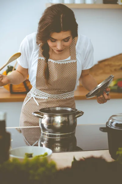 Jovem morena cozinhando sopa na cozinha. Dona de casa segurando colher de madeira na mão. Conceito de alimentação e saúde — Fotografia de Stock