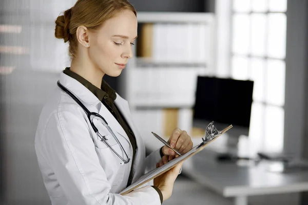 Friendly smiling female doctor using clipboard in clinic. Portrait of friendly physician woman at work. Medical service in hospital. Medicine concept