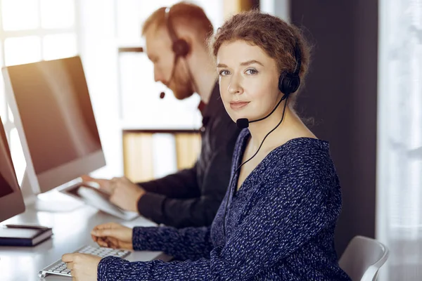 Femme d'affaires parlant par casque tout en étant assis avec un collègue à barbe rouge dans un bureau moderne ensoleillé. Groupe de personnes diversifiées dans le centre d'appels — Photo