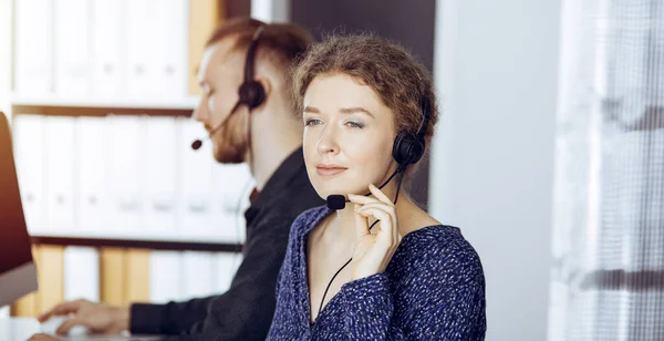 Femme d'affaires parlant par casque tout en étant assis avec un collègue à barbe rouge dans un bureau moderne ensoleillé. Groupe de personnes diversifiées dans le centre d'appels — Photo