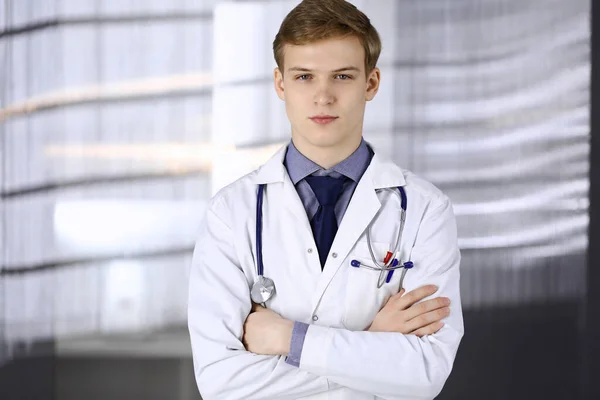 Joven médico de pie con los brazos cruzados en la clínica cerca de su lugar de trabajo. Médico rubio en el trabajo. Servicio médico perfecto en el hospital. Concepto de medicina —  Fotos de Stock