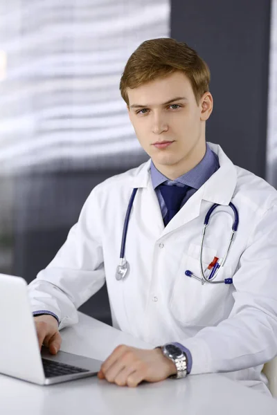 Man dokter zit en werkt met laptop in de kliniek op zijn werkplek. Blond arts op het werk. Perfecte medische dienst, medisch concept — Stockfoto