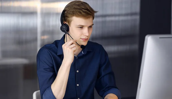 Junger blonder Geschäftsmann mit Headset und Computer bei der Arbeit. Start-up-Unternehmen bedeutet, hart zu arbeiten und keine Zeit für Erfolg zu haben — Stockfoto