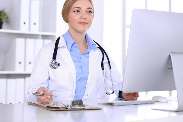 Mujer joven médico en el trabajo en el hospital mirando monitor de escritorio PC. El médico controla los registros de la historia de la medicación y los resultados del examen. Concepto de medicina y salud — Foto de Stock