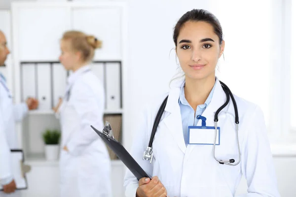 Klinikte arka planda meslektaşları olan bir kadın doktor. İspanyol ya da Latin Amerikalı tıp personeli — Stok fotoğraf