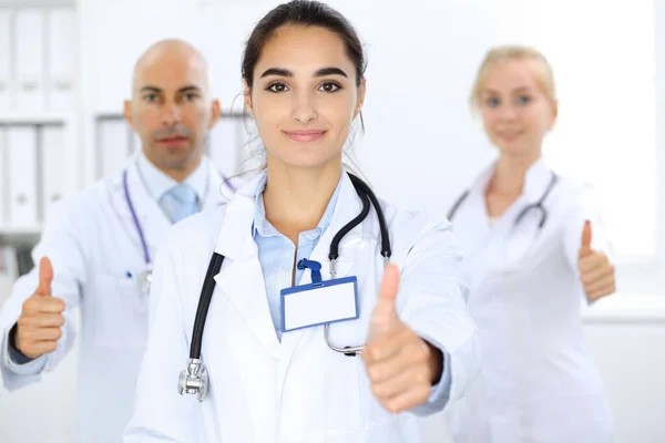 Doctor woman showing OK in clinic with colleagues at the background. Hispanic or latin american staff in medicine — Stock Photo, Image