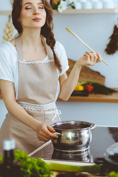 Jeune brune femme cuisine soupe dans la cuisine. Femme au foyer tenant une cuillère en bois dans sa main. Alimentation et santé concept — Photo