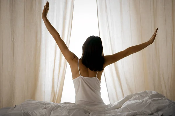 Woman stretching hands in bed after wake up, sun flare . Brunette entering a day happy and relaxed after good night sleep and back view. Concept of a new day and joyful weekend