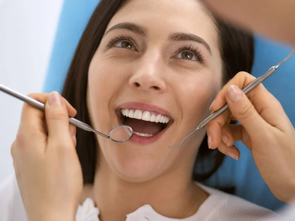 Smiling brunette woman being examined by dentist at dental clinic. Hands of a doctor holding dental instruments near patients mouth. Healthy teeth and medicine concept — Stock Photo, Image