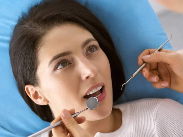 Lachende brunette vrouw wordt onderzocht door tandarts in de tandheelkundige kliniek. Handen van een arts met tandheelkundige instrumenten in de buurt van de mond van patiënten. Gezonde tanden en geneeskundeconcept — Stockfoto