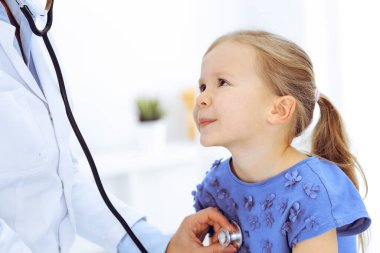 Doktor steteskopla küçük bir kızı muayene ediyor. Her zamanki tıbbi muayenede gülümseyen çocuk hasta. Tıp ve sağlık hizmetleri kavramları