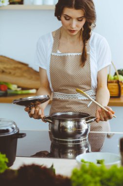 Genç esmer kadın mutfakta çorba pişiriyor. Ev hanımı elinde tahta kaşık tutuyor. Gıda ve sağlık konsepti