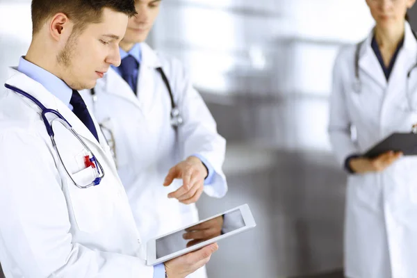 Ομάδα γιατρών ελέγχει τα ιατρικά ονόματα σε ένα τάμπλετ υπολογιστή, με μια νοσοκόμα με ένα πρόχειρο στο παρασκήνιο, στέκεται μαζί σε ένα γραφείο νοσοκομείου. Ιατροί έτοιμοι να εξετάσουν και να βοηθήσουν ασθενείς — Φωτογραφία Αρχείου