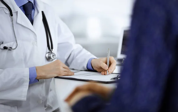 Un médico está escribiendo una receta médica para su paciente, mientras está sentado junto al escritorio en el gabinete de un hospital. Médico que usa portapapeles para llenar los registros del historial de medicamentos — Foto de Stock