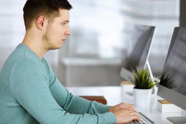 Der junge selbstbewusste Geschäftsmann sitzt in einem grünen Pullover am Schreibtisch in einem modernen Büro, während er am Computer an seinem neuen Projekt arbeitet. Kopfschuss eines männlichen Unternehmers oder IT-Spezialisten — Stockfoto