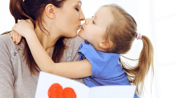 Muttertagskonzept. Die Tochter gratuliert der Mutter und überreicht ihr eine Postkarte mit rotem Herzchen. Mutter und Mädchen glücklich lächelnd und umarmt. Familienspaß und Urlaub — Stockfoto