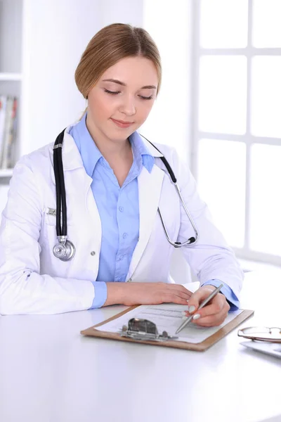 Νεαρή γυναίκα γιατρός στη δουλειά, ενώ γεμίζει το ιστορικό των φαρμάκων στο γραφείο του νοσοκομείου. Ο γιατρός ελέγχει τα αποτελέσματα των εξετάσεων. Ιατρική και υγειονομική περίθαλψη έννοια — Φωτογραφία Αρχείου