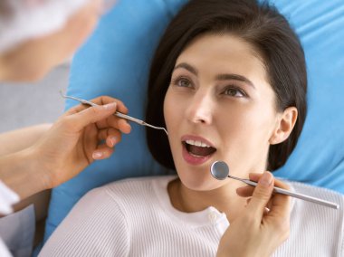 Gülümseyen esmer kadın diş hekimi tarafından muayene ediliyor. Hastanın ağzının yanında diş aletleri tutan bir doktorun elleri. Sağlıklı diş ve ilaç konsepti