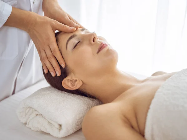 Hermosa mujer morena disfrutando de masaje facial con los ojos cerrados. Tratamiento relajante en conceptos de medicina y centro de spa — Foto de Stock