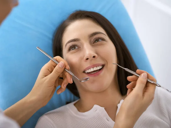 Lachende brunette vrouw wordt onderzocht door tandarts in de tandheelkundige kliniek. Handen van een arts met tandheelkundige instrumenten in de buurt van de mond van patiënten. Gezonde tanden en geneeskundeconcept — Stockfoto