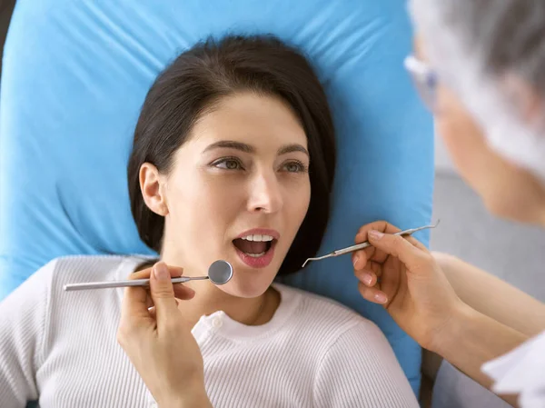 Lachende brunette vrouw wordt onderzocht door tandarts in de tandheelkundige kliniek. Handen van een arts met tandheelkundige instrumenten in de buurt van de mond van patiënten. Gezonde tanden en geneeskundeconcept — Stockfoto