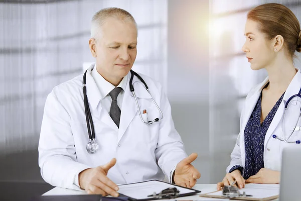 Il medico anziano e la sua collega discutono dell'attuale terapia della malattia mentre sono seduti sul posto di lavoro in una clinica soleggiata. Lavoro di squadra in medicina — Foto Stock