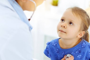 Güneşli klinikte bir çocuğu steteskopla muayene eden bir doktor. Mavi elbiseli mutlu gülümseyen kız hasta her zamanki sağlık kontrolünde.