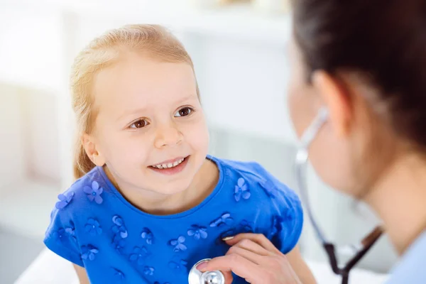 Doutor examinando uma criança por estetoscópio na clínica ensolarada. Feliz sorrindo paciente menina vestida de vestido azul é na inspeção médica habitual — Fotografia de Stock