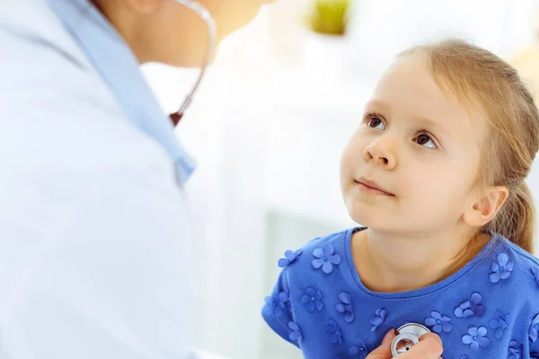 Γιατρός εξετάζει ένα παιδί με στηθοσκόπιο σε ηλιόλουστη κλινική. Ευτυχισμένο χαμογελαστό κορίτσι ασθενής ντυμένος με μπλε φόρεμα είναι στη συνήθη ιατρική επιθεώρηση — Φωτογραφία Αρχείου