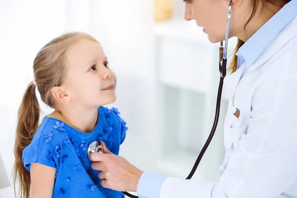 Arzt untersucht ein Kind mit Stethoskop in sonniger Klinik. Glücklich lächelnde Patientin im blauen Kleid bei der üblichen ärztlichen Untersuchung — Stockfoto