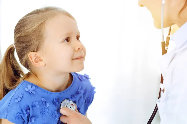 Arzt untersucht ein Kind mit Stethoskop in sonniger Klinik. Glücklich lächelnde Patientin im blauen Kleid bei der üblichen ärztlichen Untersuchung — Stockfoto