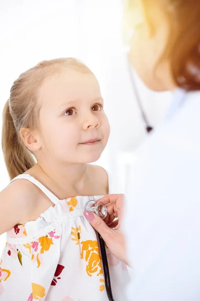 Médico examinando a un niño por estetoscopio en una clínica soleada. Feliz niña sonriente paciente vestida con vestido de color brillante es en la inspección médica habitual — Foto de Stock
